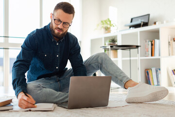 A young man sits cross-legged on the floor, focused on his studies. He writes notes in a notebook while using a laptop in a well-lit, modern living space surrounded by shelves.