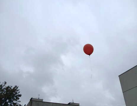 Single red balloon floating high in cloudy sky above treetops creating lonely dreamy atmosphere
