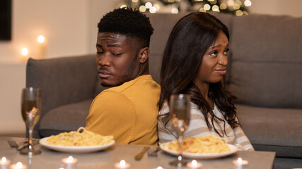 A couple shows signs of distress while sitting back-to-back on a couch after a disagreement during their Valentine's Day dinner at home. The romantic setting contrasts their mood.