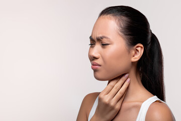 Unhappy young Asian woman shows discomfort from sore throat as she touches her neck. She is...