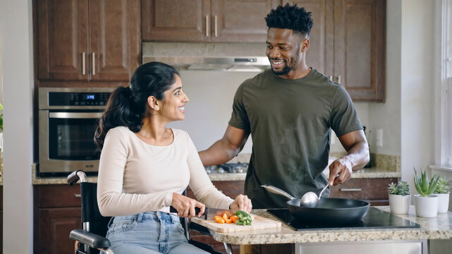 Happy interracial couple looking at each other while cooking together in the kitchen, with the woman in a wheelchair - Powered by Adobe