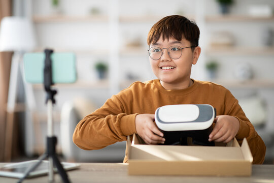 Teenager sits at a table, smiling as he unpacks VR glasses from a box. He is streaming the process on his cellphone, sharing the experience with his followers in a cozy home setting.
