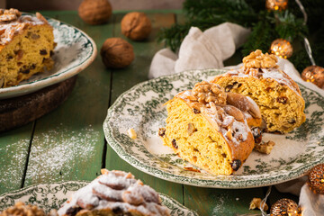 Close-up of festive nut bread slice on green floral plate, topped with sugar, almonds, walnuts, on a green table with Christmas ornaments. Bolo Rainha, a Portuguese Christmas dessert.