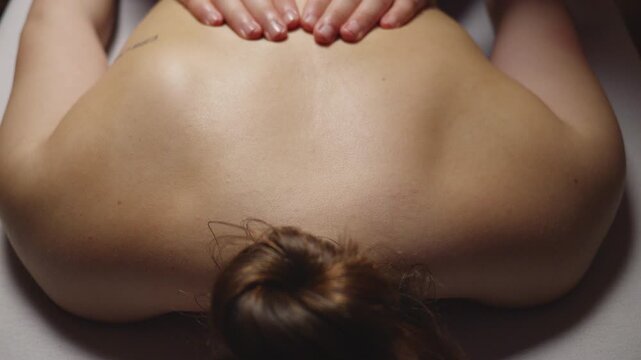 A woman lies face down on a massage table, receiving a back massage. The masseuse's hands are on her lower back, suggesting relaxation and therapeutic care in a serene spa setting.