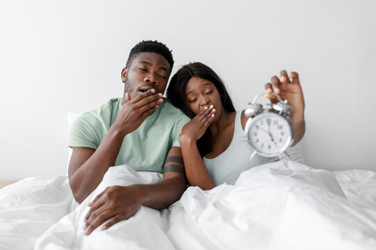 A couple sits in bed looking tired and yawning, one of them holds a clock showing early morning time. The room is bright and simply decorated, suggesting a peaceful start to the day.