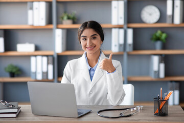 A cheerful Indian female doctor in a white coat sits at her desk in a clinic office. She is working...