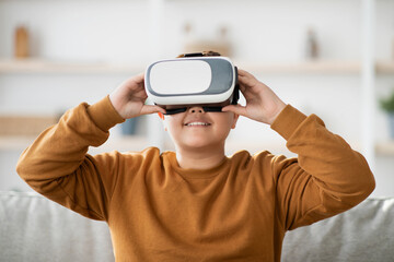 A cheerful overweight teen boy is wearing VR glasses while sitting on a comfortable couch in his living room.
