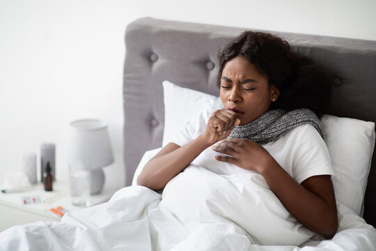 A young African American woman sits in bed with a scarf around her neck. She is coughing and appears ill at home during the pandemic, trying to rest and recover alone.