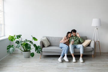 A young Asian couple sits together on a couch, happily waving at their smartphone camera during a...