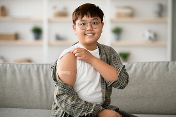 A cute plump Korean child with glasses sits on a couch at home, smiling at the camera and...
