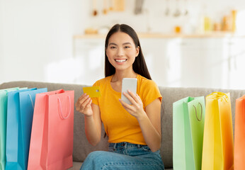 Positive asian woman sits on a sofa surrounded by vibrant shopping bags. She smiles at the camera...