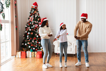 Cheerful black girl dances joyfully with her parents in a warm living room. The family wears Santa...