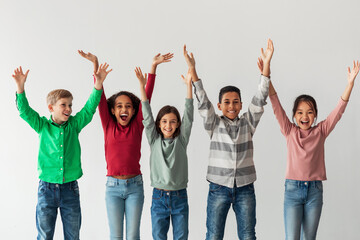 A group of preteen kids stand together with their hands raised in joy. They are smiling widely,...