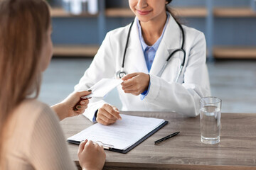 A smiling Indian female doctor in a white coat accepts payment from a client in a clinic office. The setting is professional, illustrating a typical interaction in a medical facility.