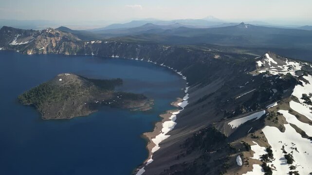 Epic cinematic aerial drone view of Crater Lake National Park, Oregon, featuring Wizard Island, snow-covered volcanic cliffs, and deep blue caldera water under clear summer sky
