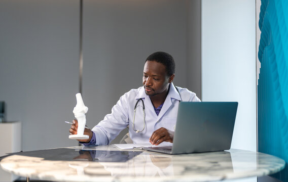 African doctor with stethoscope with medical report while working on laptop and medical documents in hospital, concept of healthcare, telemedicine, diagnosis, electronic records and healthtech