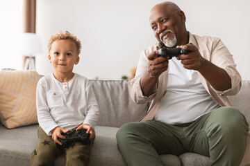 A happy grandfather and his young grandson are sitting on the sofa, playing video games together....