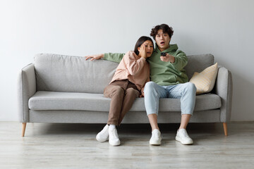 A young Asian couple sits on a couch at home, watching a horror movie on TV. Both are scared and...