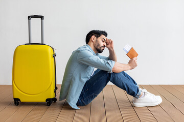 A man sits on the floor next to a yellow suitcase, visibly worried as he checks his travel...