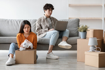 A frustrated young Asian woman unpacks a carton box alone on moving day. Her boyfriend stays on the...