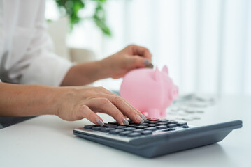 Person saving money inserting coin into pink piggy bank while calculating budget, personal finance, budgeting and financial planning at home with calculator, financial goal management, cost control