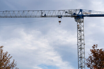 A large blue and silver construction tower crane extends across a cloudy sky, framed by autumn trees below.