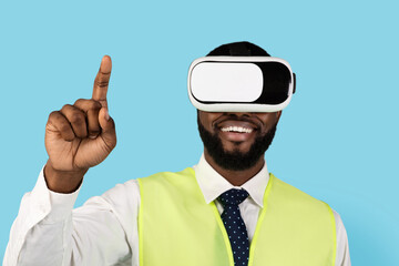 Black Builder Man Using VR Glasses And Touching Air With Finger, Happy African American Male Constructor Experiencing Virtual Reality, Enjoying Video Games While Standing On Blue Background