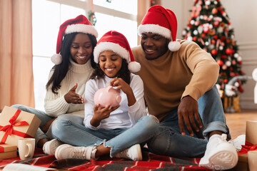 A joyful African American family sits on the floor at home, putting coins into a piggy bank. The...