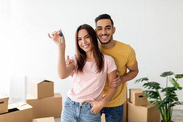Excited couple holds keys and smiles joyfully in their new apartment. They stand amidst unpacked boxes, ready to start a new chapter in their lives.