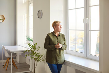 Portrait of thoughtful mature woman standing near window, gazing outside with reflective...