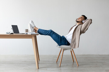 Fototapeta premium An Asian man is comfortably seated in a chair, wearing headphones and listening to music. He leans back with a smile, legs crossed on the table, enjoying a moment of relaxation at his desk.