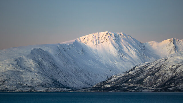 Sunlit snowy mountain above Arctic fjord in Tromsø, Norway at sunrise or sunset