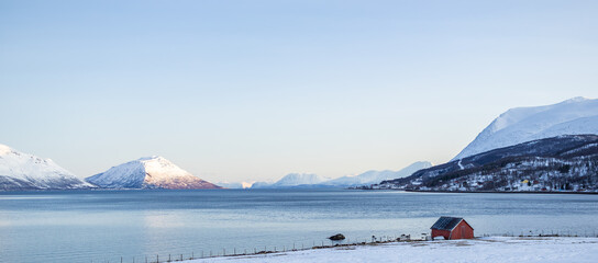A peaceful Arctic winter landscape near Tromsø, Norway, showing a calm fjord surrounded by snow-covered mountains under a clear blue sky. A small red cabin sits alone by the snowy shoreline.