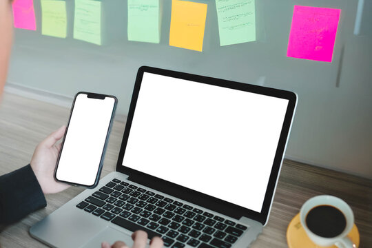 A person multitasks with a blank laptop and smartphone in an office. A glass board with colorful sticky notes is in the background, ideal for brainstorming or project planning concepts.