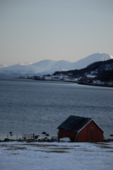Red cabin by snowy fjord with mountain backdrop in Tromsø, Norway. Arctic winter landscape.