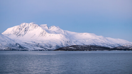 A stunning winter view of a fjord near Tromsø, Norway, featuring calm blue water and towering snow-covered mountains bathed in soft Arctic light.