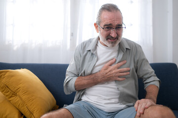Fototapeta premium Elderly man sitting on a sofa and holding his chest in pain