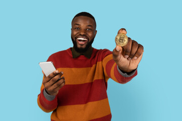 Online Trading. Excited Young Black Man Holding Golden Bitcoin And Smartphone, Cheerful African American Male Earning Cryptocurrency While Standing Over Blue Studio Background, Closeup