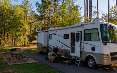 An RV is parked at a quiet campsite surrounded by tall trees. The scene is calm with a picnic table and chairs set up outside the vehicle.