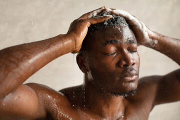 A young African American man stands in a modern bathroom, relaxing under falling water. He washes his hair, enjoying a soothing shower experience. The atmosphere is calm and refreshing.