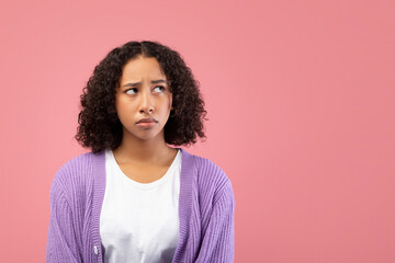 A young black woman looks away thoughtfully in a pink studio setting. She appears to be upset or doubtful, lost in her own thoughts. Her expression reflects deep contemplation and concern.
