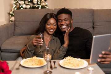 Happy black couple enjoys a cozy Valentine's Day at home. They take a selfie on a tablet while sharing dinner and watching romantic movies, creating lasting memories together.