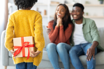 A young child hides a gift behind their back while two smiling parents watch eagerly. The scene...