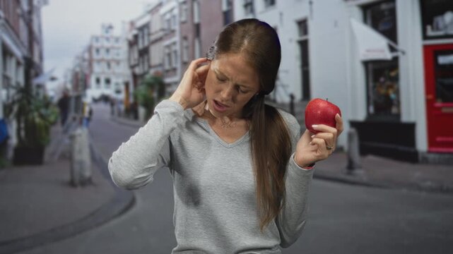 Young hispanic woman scratches neck while holding a red apple on a street; discomfort food dilemma moment.
