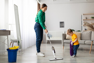 A woman uses a mop to clean the living room floor while her young child plays nearby, engaged with a toy. The scene reflects a caring home atmosphere and daily chores.