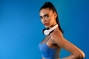 Fit female stands confidently on a blue background wearing sportswear and wireless headphones. She...