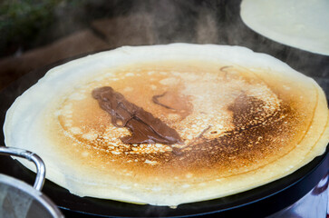 Large, thin golden crepe is cooking on a griddle, with a dollop of melted chocolate hazelnut spread in the center. Steam rises from the hot dessert