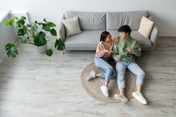 A young Asian couple smiles as they sit on the floor at home, looking at a smartphone together....