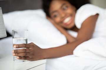 A smiling young woman reaches for a glass of water at her bedside after waking up. It is morning, and she is relaxing in bed, promoting hydration and wellness.
