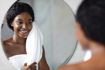 A beautiful african american woman smiles as she enjoys her morning beauty routine after showering. She stands in front of a mirror, gently touching her face with a towel.